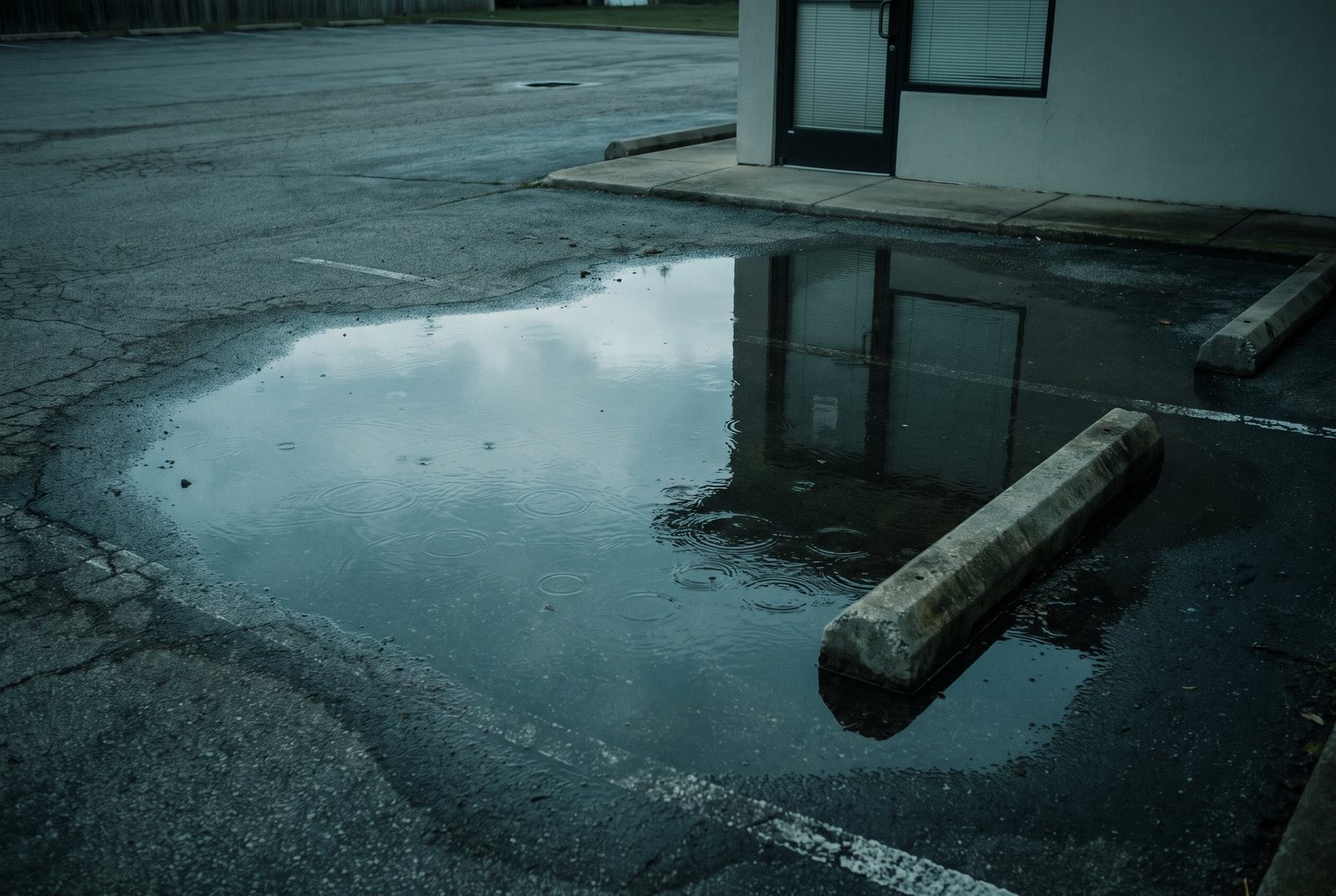 A rain puddle in an empty parking lot reflecting a closed office door with drawn blinds.