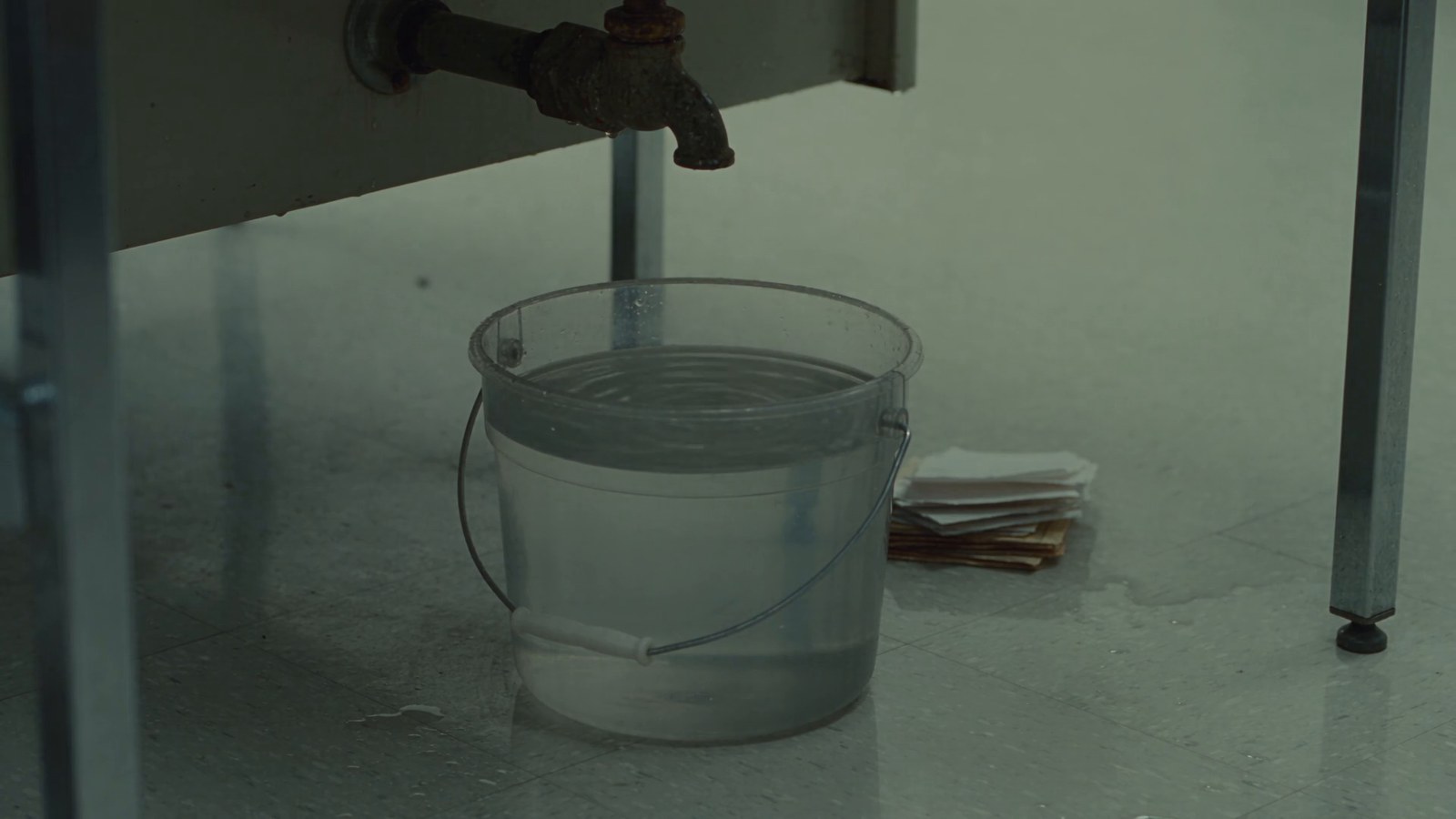 A pipe joint under a bare desk drips into a nearly full plastic bucket beside a soggy stack of paper.